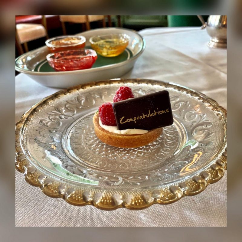 Mini pastry case filled with cream, 3 raspberries and a dark chocolate sign with Congratulations written on it in gold